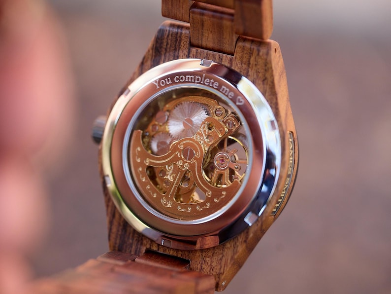 Puede incluir: Un reloj de pulsera de madera con una parte posterior transparente que revela el intrincado movimiento mec&aacute;nico de color dorado. La esfera del reloj tiene la inscripci&oacute;n "You complete me" con un s&iacute;mbolo de coraz&oacute;n.