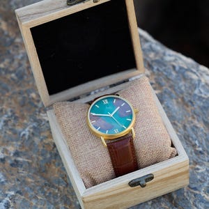 Puede incluir: Un reloj dorado con esfera verde azulado y morada y correa de cuero marr&oacute;n descansa en una caja de madera. La esfera del reloj presenta n&uacute;meros romanos y agujas blancas. La caja est&aacute; forrada con tela beige.