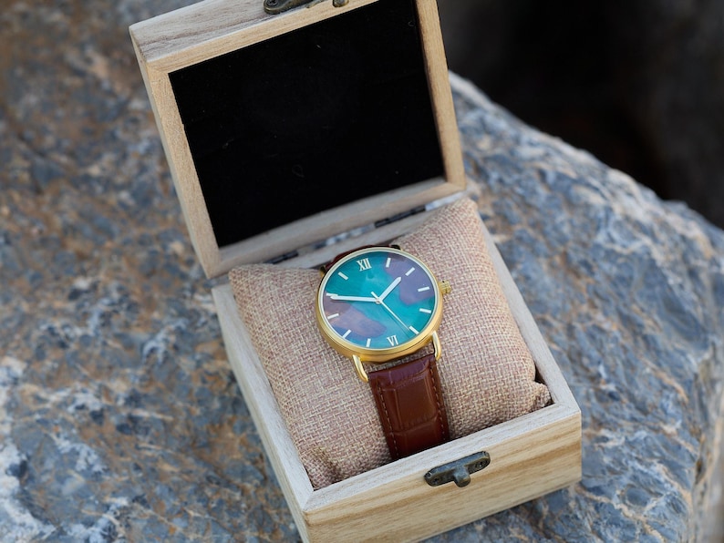 Puede incluir: Un reloj dorado con esfera verde azulado y morada y correa de cuero marr&oacute;n descansa en una caja de madera. La esfera del reloj presenta n&uacute;meros romanos y agujas blancas. La caja est&aacute; forrada con tela beige.