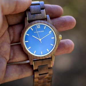 May include: Wooden wristwatch with a blue face and gold-colored accents. The watch features a dark brown wooden band and a round face with gold hands and markers. The watch face has the word "WOODPECKER" printed on it.