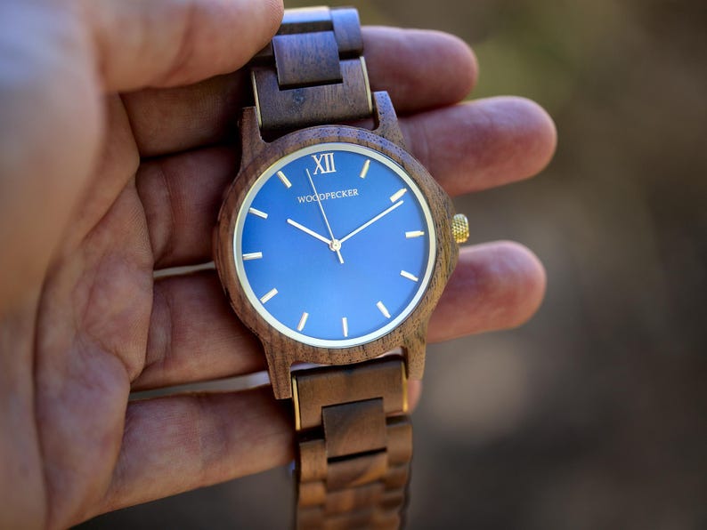 May include: Wooden wristwatch with a blue face and gold-colored accents. The watch features a dark brown wooden band and a round face with gold hands and markers. The watch face has the word "WOODPECKER" printed on it.