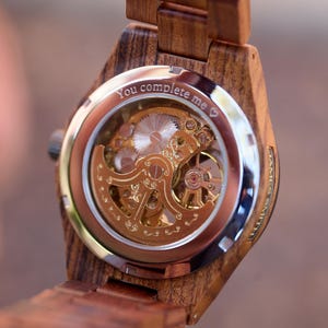 May include: Close-up of a wooden wristwatch with a visible mechanical movement. The watch has a brown wooden band and a silver and gold-toned face. The text "You complete me" is engraved on the watch face.