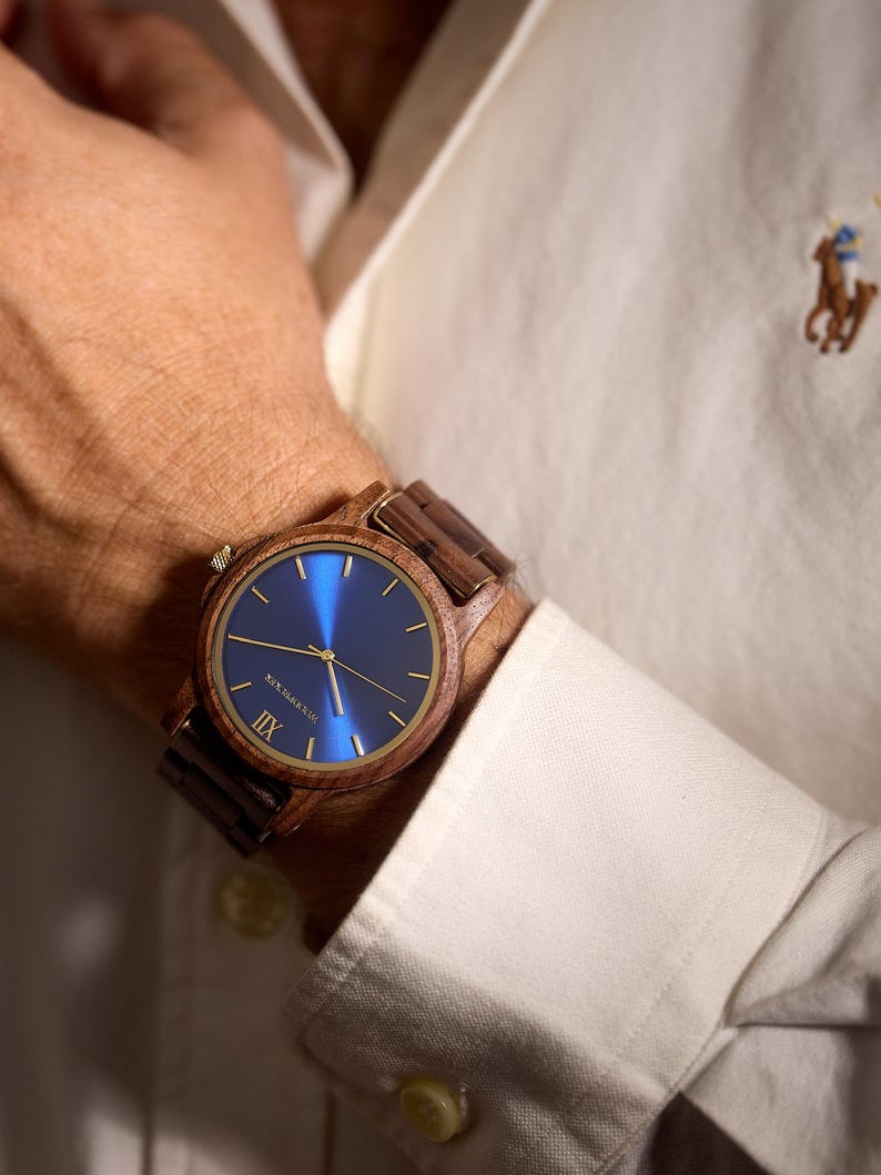 May include: A wooden wristwatch with a blue face and gold-colored hands and accents. The watch has a brown wooden band and is worn on a wrist. The watch face has the text "NEW ZEALAND" and the Roman numeral "IIX".