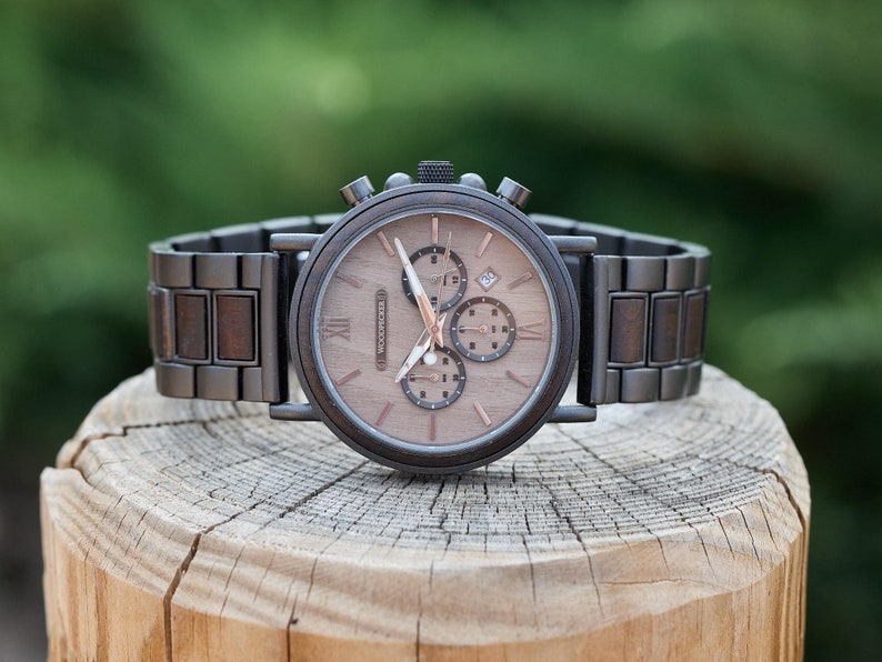 Pu&ograve; includere: Un orologio da polso in legno marrone con un quadrante cronografo e un cinturino in metallo marrone. L'orologio &egrave; su una superficie di legno.