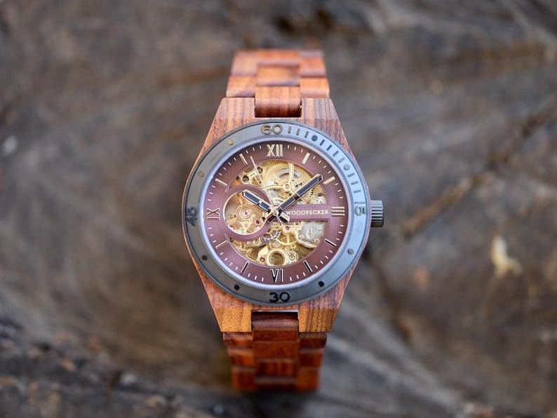 May include: A wooden wristwatch with a brown face and band. The watch face has a visible mechanical movement and Roman numeral hour markers. The watch is branded "WOODPECKER".