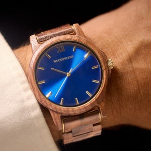 May include: Wooden wristwatch with a blue face and gold-colored hands and markers. The watch has a wooden band and a gold-colored crown. The watch face has the word "WOODPICKER" printed on it.