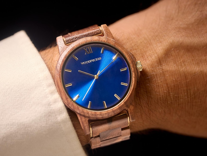 May include: Wooden wristwatch with a blue face and gold-colored hands and markers. The watch has a wooden band and a gold-colored crown. The watch face has the word "WOODPICKER" printed on it.