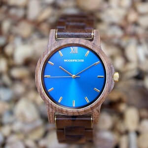 May include: A wooden wristwatch with a blue face and gold accents. The watch features a wooden band and a gold crown. The watch face has the word "WOODPECKER" printed on it. The watch is set against a blurred background of small rocks.