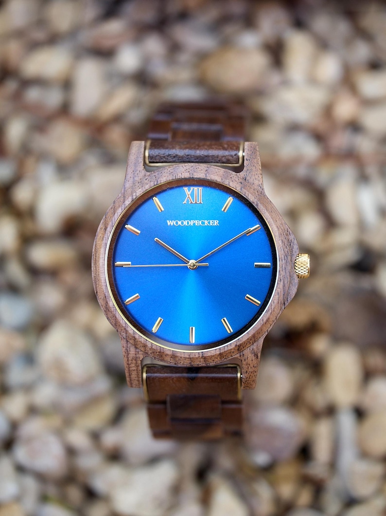 May include: A wooden wristwatch with a blue face and gold accents. The watch features a wooden band and a gold crown. The watch face has the word "WOODPECKER" printed on it. The watch is set against a blurred background of small rocks.