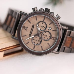 Pu&ograve; includere: Un orologio cronografo in legno marrone con cinturino in metallo nero. Il quadrante dell'orologio ha indici orari con numeri romani e una finestrella per la data alle 4. L'orologio &egrave; realizzato da Woodpecker.