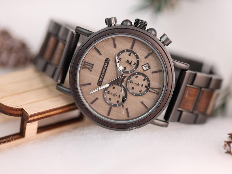 Pu&ograve; includere: Un orologio cronografo in legno marrone con cinturino in metallo nero. Il quadrante dell'orologio ha indici orari con numeri romani e una finestrella per la data alle 4. L'orologio &egrave; realizzato da Woodpecker.