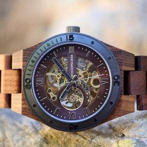 Puede incluir: Un reloj de pulsera de madera marr&oacute;n con un bisel negro y un movimiento autom&aacute;tico esqueletizado. La esfera del reloj tiene marcadores de hora en n&uacute;meros romanos y una ventana de fecha en la posici&oacute;n de las 3 en punto. La correa del reloj est&aacute; hecha de madera marr&oacute;n oscura.