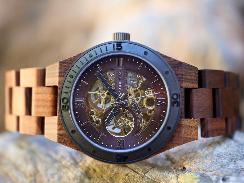 Puede incluir: Un reloj de pulsera de madera marr&oacute;n con un bisel negro y un movimiento autom&aacute;tico esqueletizado. La esfera del reloj tiene marcadores de hora en n&uacute;meros romanos y una ventana de fecha en la posici&oacute;n de las 3 en punto. La correa del reloj est&aacute; hecha de madera marr&oacute;n oscura.