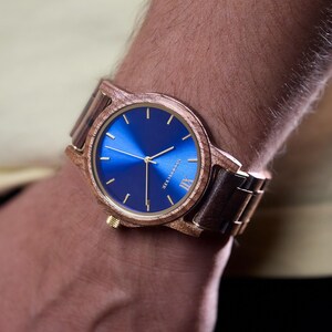 May include: A wooden wristwatch with a blue face and gold hands. The watch has a wooden bezel and a brown wooden band. The watch is on a person's wrist.