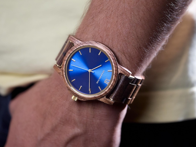 May include: A wooden wristwatch with a blue face and gold hands. The watch has a wooden bezel and a brown wooden band. The watch is on a person's wrist.