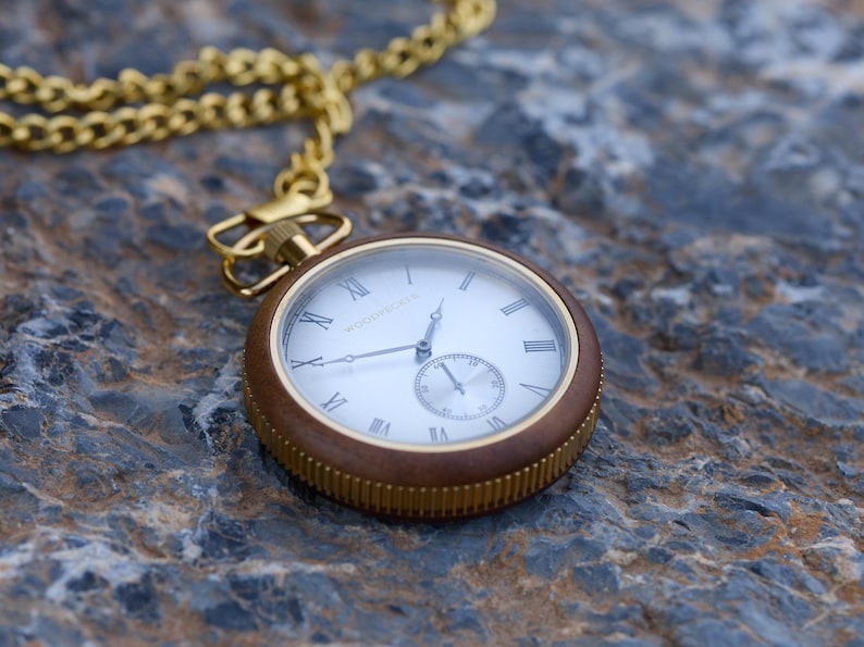 May include: A gold pocket watch with a brown wooden case and a white face with Roman numeral markings. The watch has a gold chain attached to it.