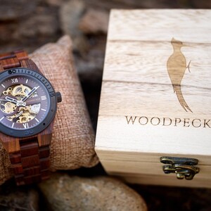 May include: A wooden wristwatch with a brown band and a dark face, showcasing the inner workings. The watch is displayed next to a wooden box with the word "WOODPECKER" and a woodpecker silhouette.