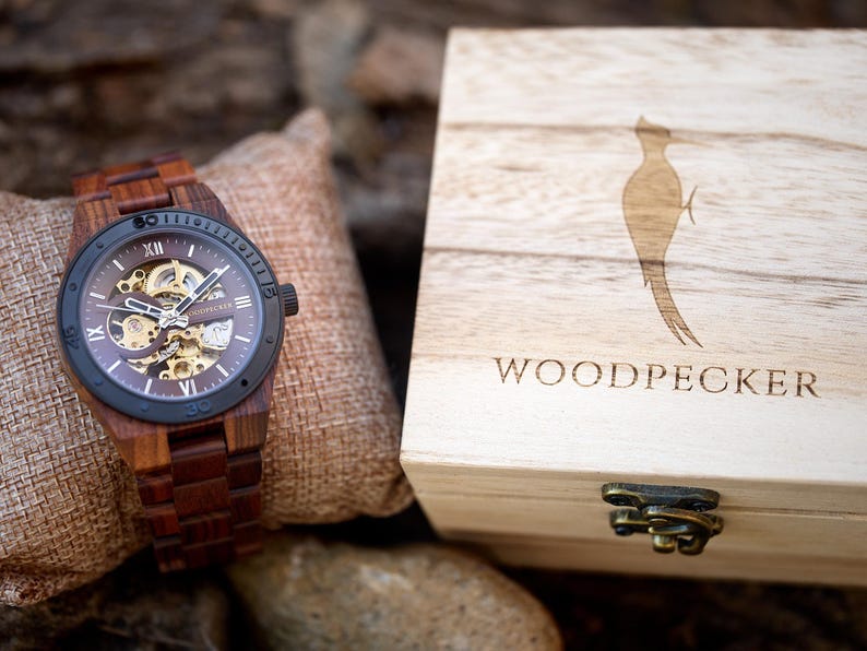 May include: A wooden wristwatch with a brown band and a dark face, showcasing the inner workings. The watch is displayed next to a wooden box with the word "WOODPECKER" and a woodpecker silhouette.