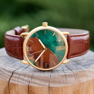 Puede incluir: Reloj dorado con correa de cuero marr&oacute;n. La esfera combina madera y resina verde, con agujas doradas y n&uacute;meros romanos. La correa es marr&oacute;n. El di&aacute;metro del reloj es de aproximadamente 4 cm.