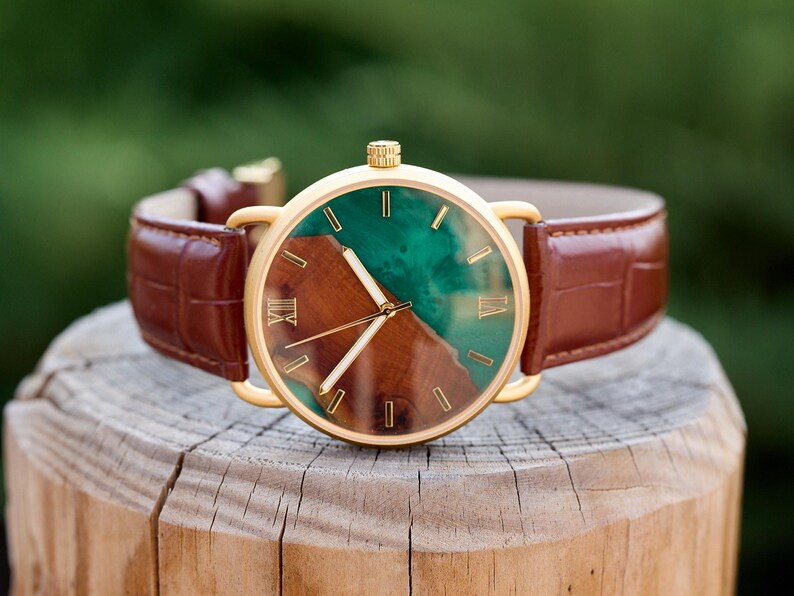 Puede incluir: Reloj dorado con correa de cuero marr&oacute;n. La esfera combina madera y resina verde, con agujas doradas y n&uacute;meros romanos. La correa es marr&oacute;n. El di&aacute;metro del reloj es de aproximadamente 4 cm.