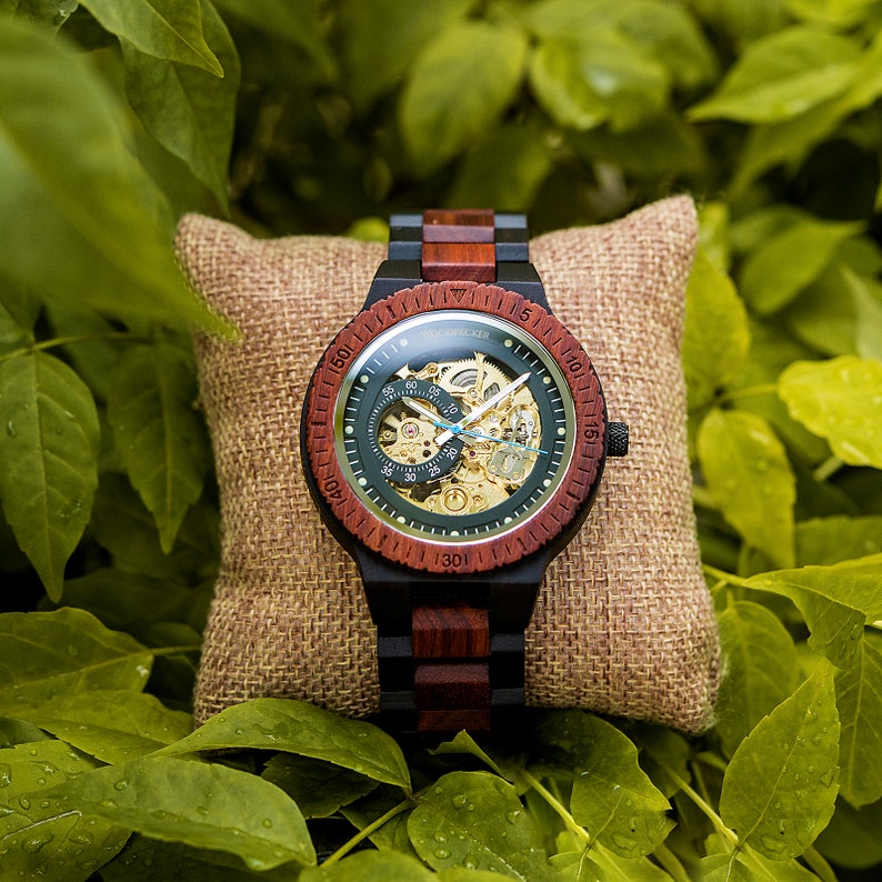 May include: A wooden wristwatch with a green face and a red and black wooden band. The watch has a skeleton design, revealing the inner workings of the watch. The watch is on a brown burlap pillow.