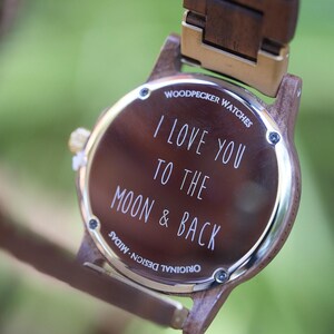 May include: A wooden wristwatch with a brown face and band. The watch face has the inscription "I LOVE YOU TO THE MOON & BACK" in white lettering. The watch is from Woodpecker Watches.