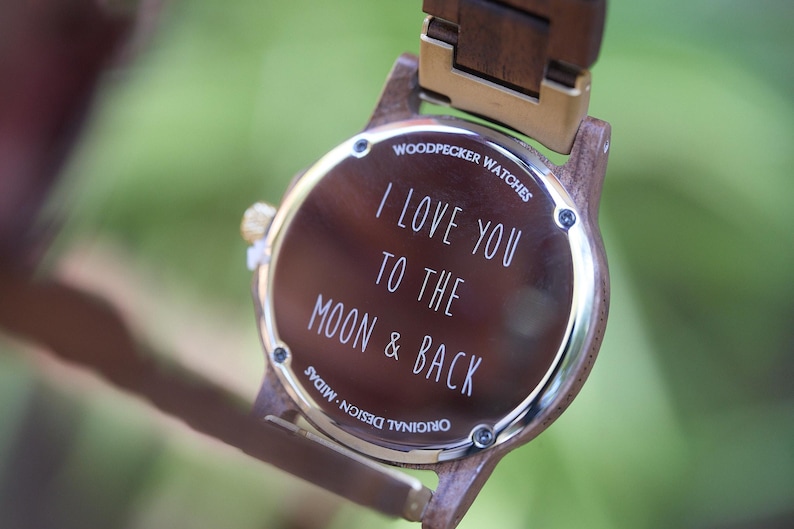 May include: A wooden wristwatch with a brown face and band. The watch face has the inscription "I LOVE YOU TO THE MOON & BACK" in white lettering. The watch is from Woodpecker Watches.