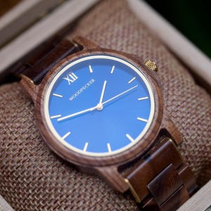 May include: Wooden wristwatch with a blue face and gold-colored hands and accents. The watch is in a wooden box with a burlap lining. The watch face has the word "WOODPECKER" printed on it.