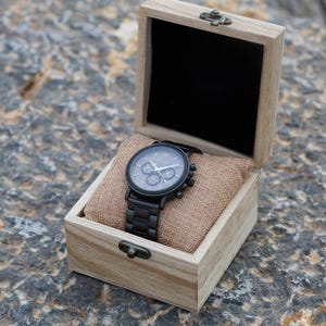Pu&ograve; includere: Un orologio da polso cronografo nero con cinturino in pelle marrone &egrave; esposto in una scatola di legno con coperchio a cerniera.