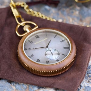 May include: A gold pocket watch with a wooden case and a white face. The watch has Roman numeral hour markers and a smaller subdial for seconds. The watch is on a brown suede pouch.