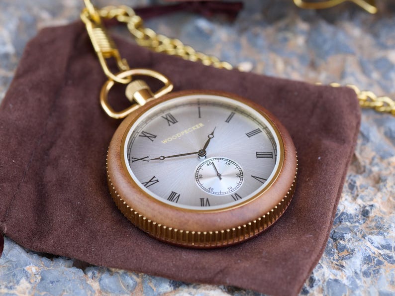 May include: A gold pocket watch with a wooden case and a white face. The watch has Roman numeral hour markers and a smaller subdial for seconds. The watch is on a brown suede pouch.