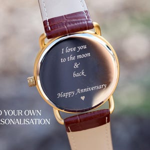Puede incluir: Reloj dorado con esfera oscura y correa de cuero. La esfera del reloj tiene la inscripci&oacute;n "I love you to the moon & back Happy Anniversary" en escritura blanca. La correa es marr&oacute;n y crema.