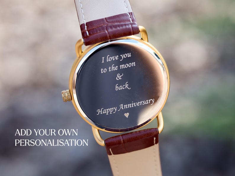Puede incluir: Reloj dorado con esfera oscura y correa de cuero. La esfera del reloj tiene la inscripci&oacute;n "I love you to the moon & back Happy Anniversary" en escritura blanca. La correa es marr&oacute;n y crema.