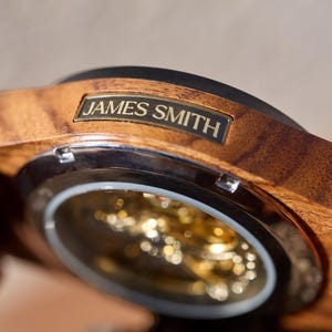 Puede incluir: Un reloj de pulsera de madera con una esfera dorada y una correa de metal. La esfera del reloj es visible a trav&eacute;s de una cubierta de vidrio transparente. El reloj tiene una inscripci&oacute;n personalizada que dice "JAMES SMITH".