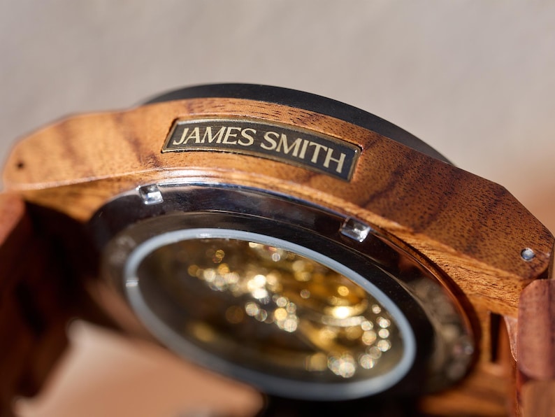 Puede incluir: Un reloj de pulsera de madera con una esfera dorada y una correa de metal. La esfera del reloj es visible a trav&eacute;s de una cubierta de vidrio transparente. El reloj tiene una inscripci&oacute;n personalizada que dice "JAMES SMITH".