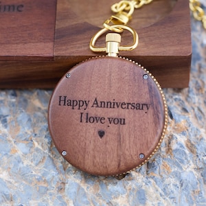 May include: A wooden pocket watch with a gold chain. The watch face is engraved with the words "Happy Anniversary I love you" and a small heart.