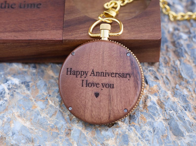 May include: A wooden pocket watch with a gold chain. The watch face is engraved with the words "Happy Anniversary I love you" and a small heart.
