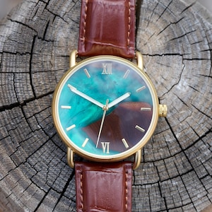 Puede incluir: Un reloj dorado con una correa de cuero marr&oacute;n. La esfera presenta un dise&ntilde;o veteado turquesa y marr&oacute;n, manecillas doradas y n&uacute;meros romanos. El reloj se encuentra sobre una superficie de madera envejecida.