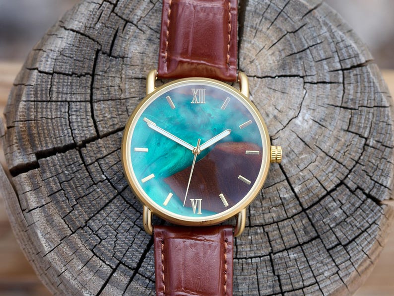 Puede incluir: Un reloj dorado con una correa de cuero marr&oacute;n. La esfera presenta un dise&ntilde;o veteado turquesa y marr&oacute;n, manecillas doradas y n&uacute;meros romanos. El reloj se encuentra sobre una superficie de madera envejecida.