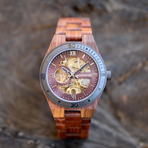 Puede incluir: Un reloj de pulsera de madera marr&oacute;n con un movimiento autom&aacute;tico esqueletizado. La esfera del reloj tiene marcadores de hora romanos negros y una aguja de segundos negra. La correa del reloj est&aacute; hecha de madera marr&oacute;n oscura.