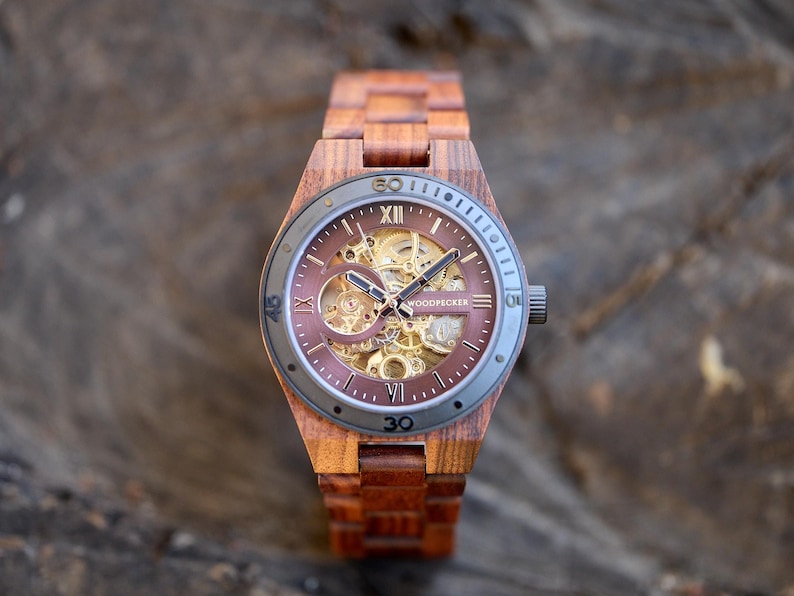 Puede incluir: Un reloj de pulsera de madera marr&oacute;n con un movimiento autom&aacute;tico esqueletizado. La esfera del reloj tiene marcadores de hora romanos negros y una aguja de segundos negra. La correa del reloj est&aacute; hecha de madera marr&oacute;n oscura.