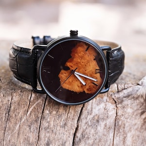 Puede incluir: Un reloj de pulsera negro con una esfera marr&oacute;n oscuro con un dise&ntilde;o de veta de madera marr&oacute;n claro. El reloj tiene manecillas negras y marcadores de hora con n&uacute;meros romanos. Tiene una correa de cuero negro con estampado de cocodrilo, sobre madera envejecida.