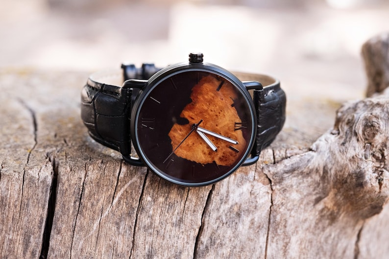 Puede incluir: Un reloj de pulsera negro con una esfera marr&oacute;n oscuro con un dise&ntilde;o de veta de madera marr&oacute;n claro. El reloj tiene manecillas negras y marcadores de hora con n&uacute;meros romanos. Tiene una correa de cuero negro con estampado de cocodrilo, sobre madera envejecida.