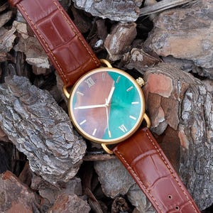 Puede incluir: Un reloj dorado con una correa de cuero marr&oacute;n. La esfera del reloj est&aacute; dividida en secciones marrones y turquesas, con n&uacute;meros romanos. El reloj se muestra sobre un lecho de astillas de madera marrones y grises.