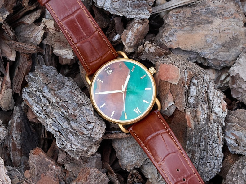Puede incluir: Un reloj dorado con una correa de cuero marr&oacute;n. La esfera del reloj est&aacute; dividida en secciones marrones y turquesas, con n&uacute;meros romanos. El reloj se muestra sobre un lecho de astillas de madera marrones y grises.