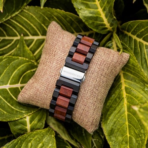 May include: A black and brown wooden watch band with a silver buckle. The band is on a brown fabric cushion and says "WOODPECKER" on the buckle.