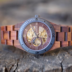 May include: A wooden wristwatch with a brown face and band. The watch face has a visible mechanical movement and Roman numeral hour markers. The watch is made of wood and has a dark gray bezel. The brand name "WOODPECKER" is visible on the watch face.