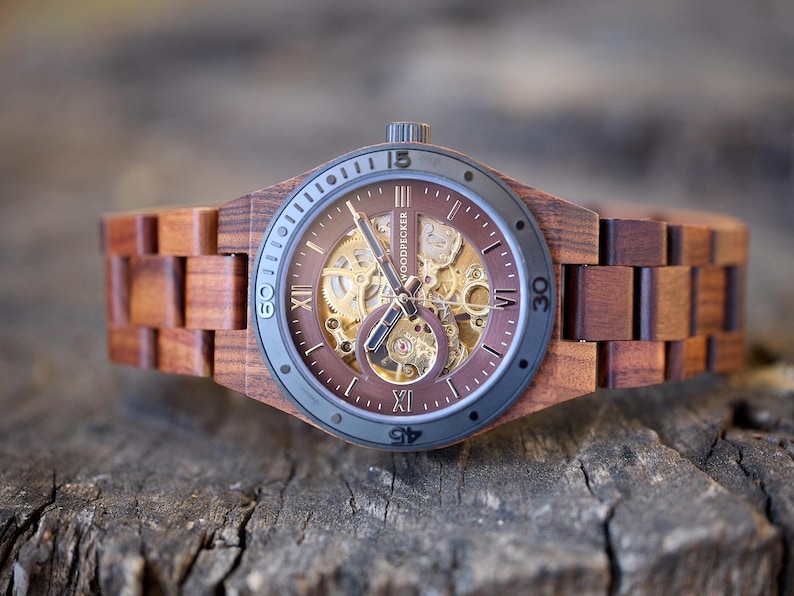 May include: A wooden wristwatch with a brown face and band. The watch face has a visible mechanical movement and Roman numeral hour markers. The watch is made of wood and has a dark gray bezel. The brand name "WOODPECKER" is visible on the watch face.