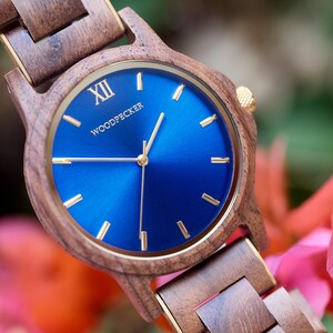 May include: A wooden wristwatch with a blue face and gold accents. The watch face displays the word "WOODPECKER" and the Roman numeral "XII". The watch band and case are made of wood.