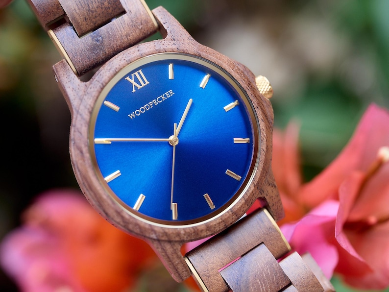 May include: A wooden wristwatch with a blue face and gold accents. The watch face displays the word "WOODPECKER" and the Roman numeral "XII". The watch band and case are made of wood.